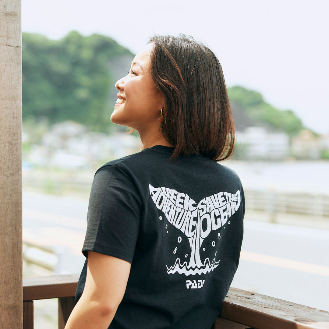 a model wearing a black t shirt with white whale tail design written 'Seek Adventure, Save the Ocean'