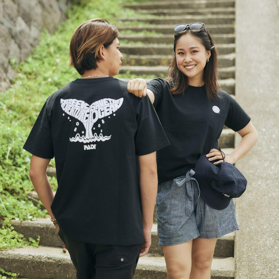 Two models wearing a black t shirt with white whale tail design written 'Seek Adventure, Save the Ocean'