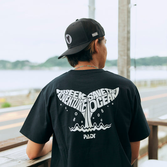 a model wearing a black t shirt with white whale tail design written 'Seek Adventure, Save the Ocean'